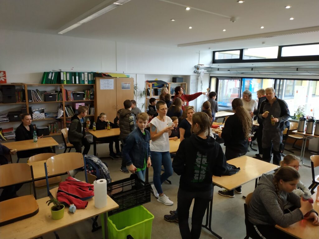 Viele Schüler stehen und sitzen in einem Klassenraum, unterhalten sich und arbeiten.