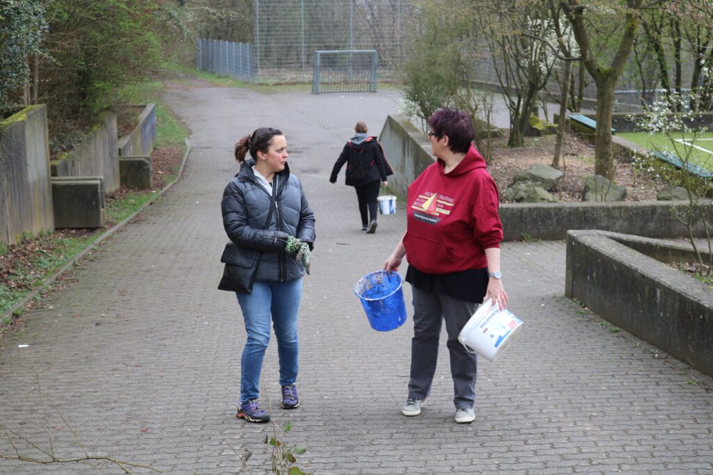 Zwei Frauen gehen mit Eimern einen Weg entlang, eine weitere Person ist im Hintergrund.