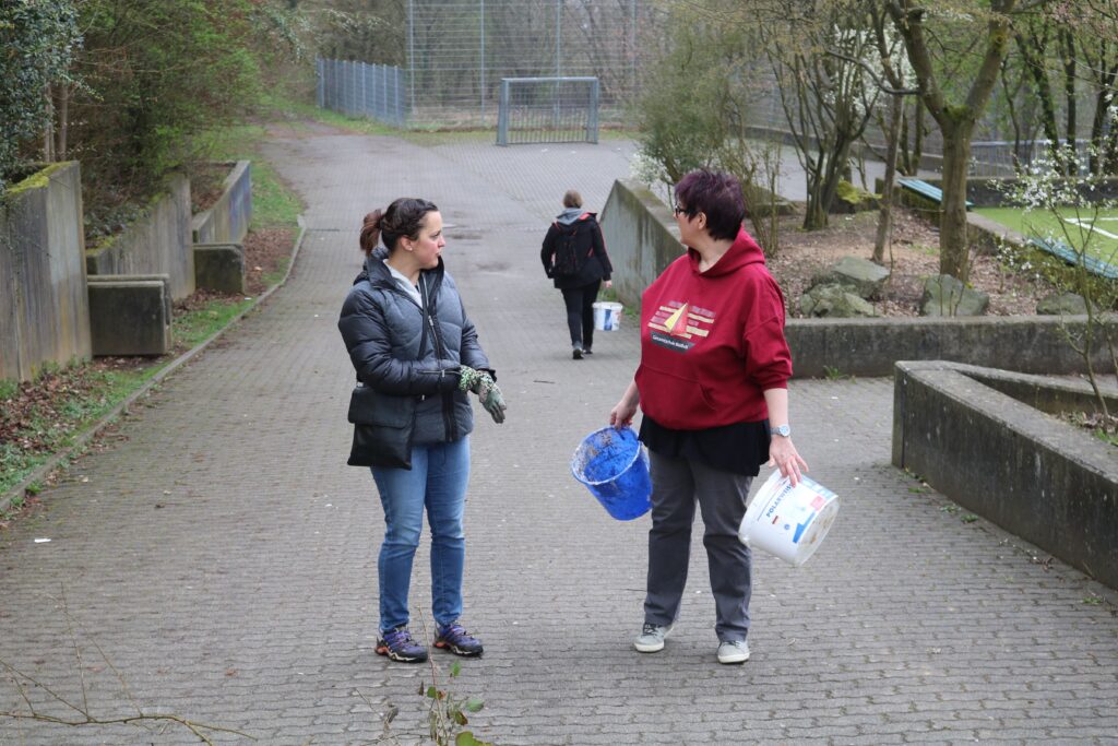 Zwei Frauen stehen auf einem Weg mit Eimern, eine Person geht im Hintergrund.
