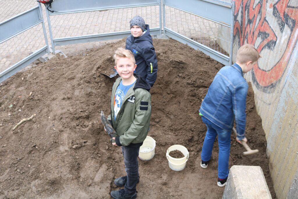 Drei Kinder graben in einem Sandhaufen und lächeln in die Kamera.