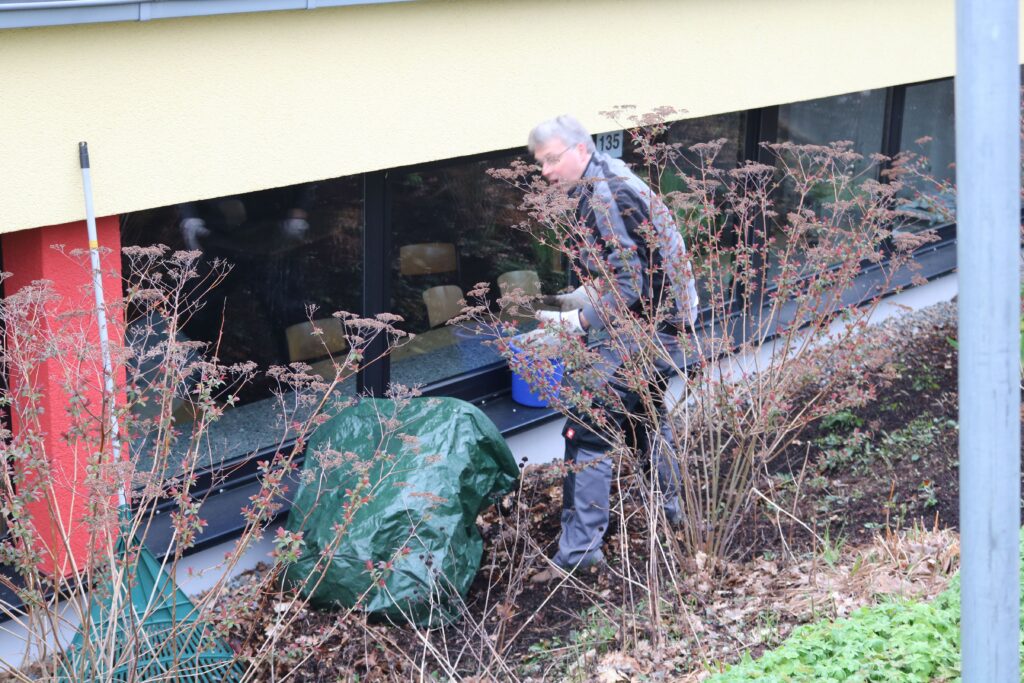 Mann arbeitet im Garten neben einem Gebäude und bewegt Pflanzen oder Materialien.