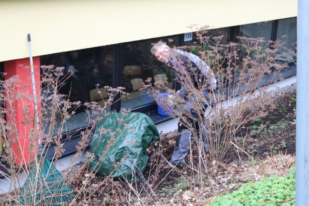 Person arbeitet im Garten neben einem Gebäude. Sträucher und grüne Abdeckung sichtbar.