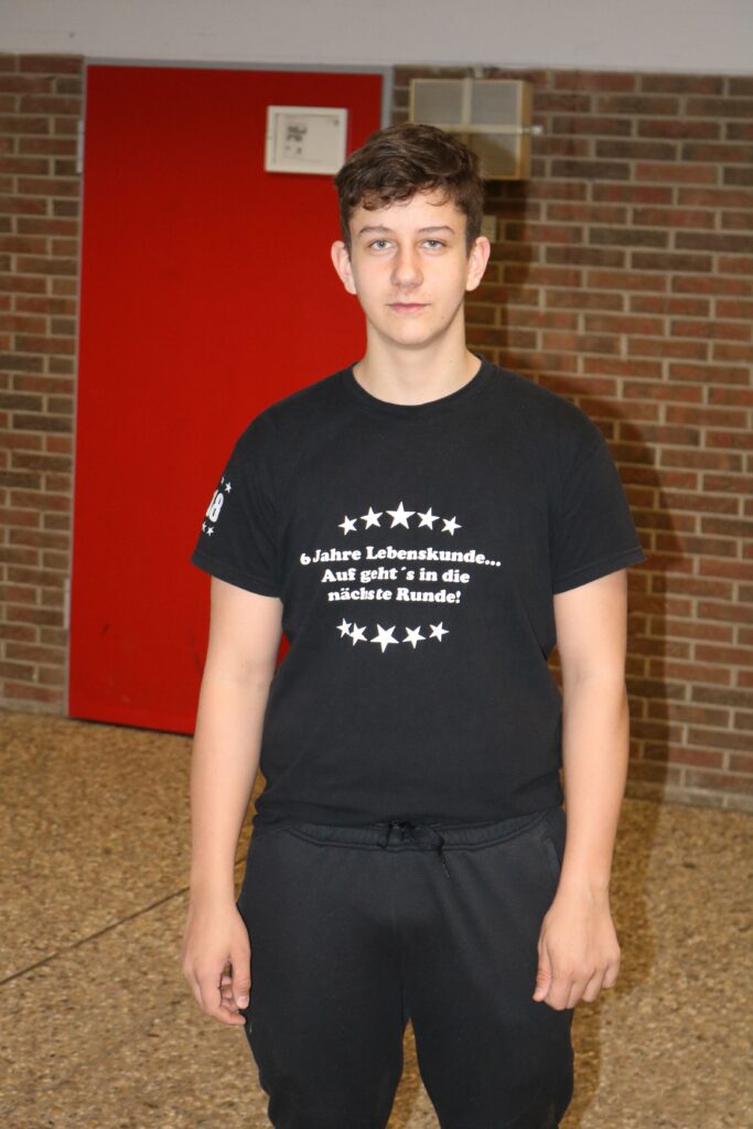 Schüler steht vor einer roten Tür in einem Flur, trägt ein schwarzes T-Shirt.