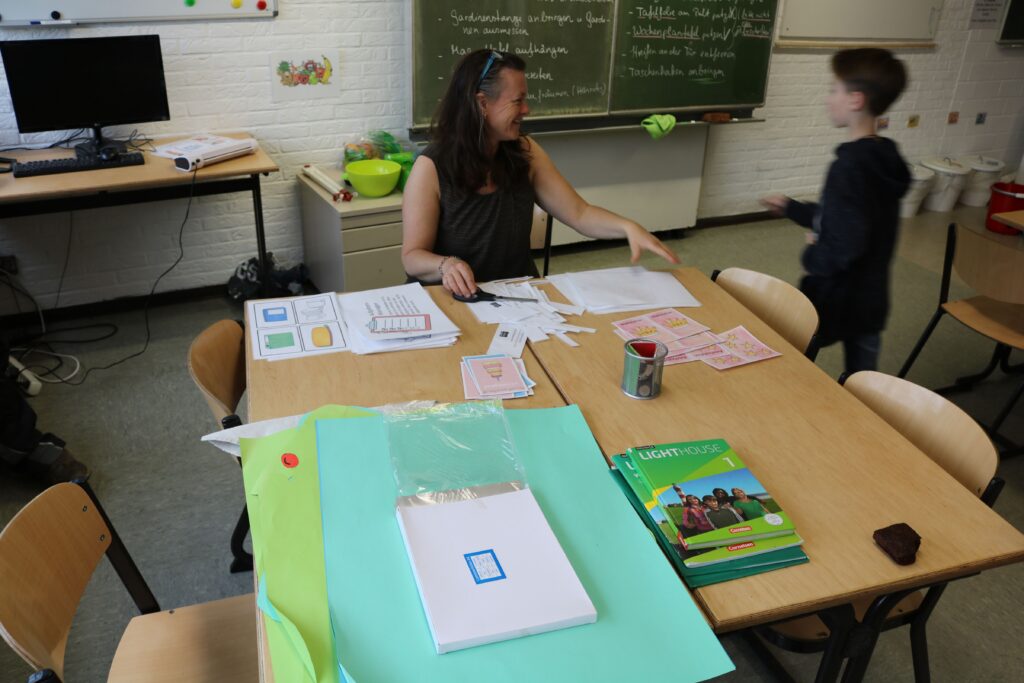 Lehrerin und Schüler arbeiten an einem Tisch mit verschiedenen Materialien in einem Klassenzimmer.