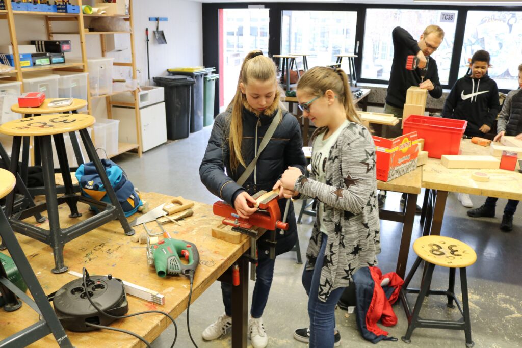 Schülerinnen arbeiten an einem Tisch im Werkraum mit Werkzeugen und Materialien.