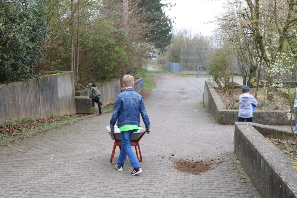 Kinder helfen im Freien, ein Junge schiebt eine Schubkarre, andere sind aktiv.