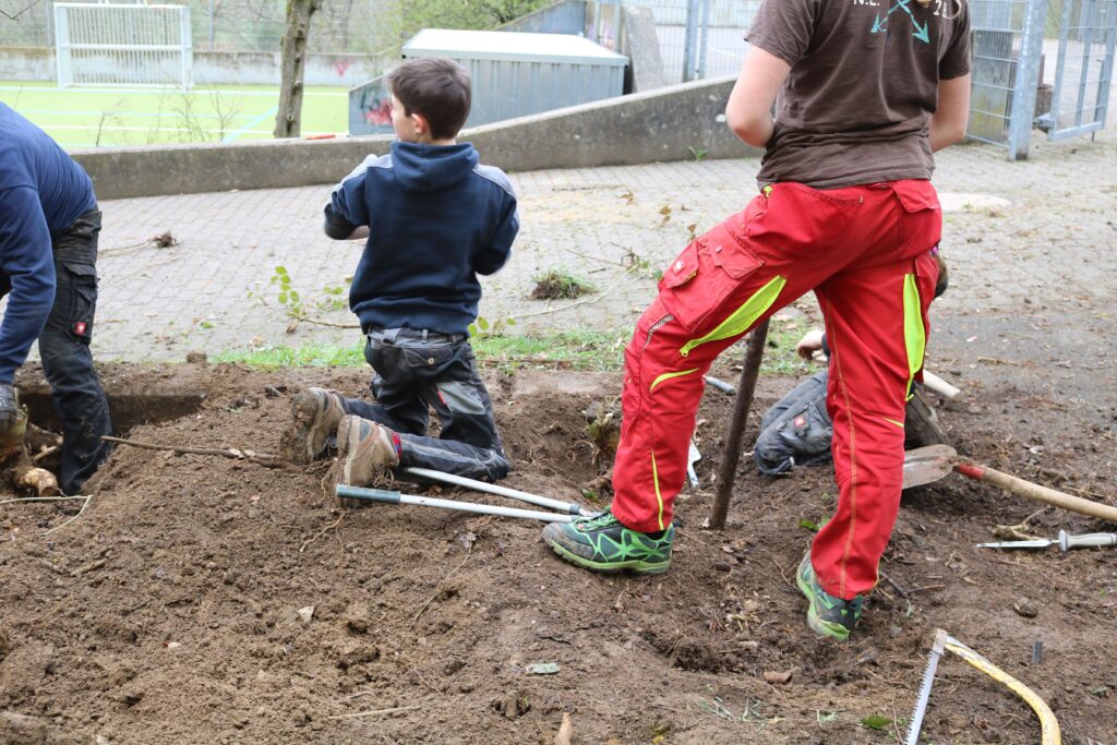 Kinder buddeln auf einem Schulhof, Erde und Werkzeuge sind sichtbar.