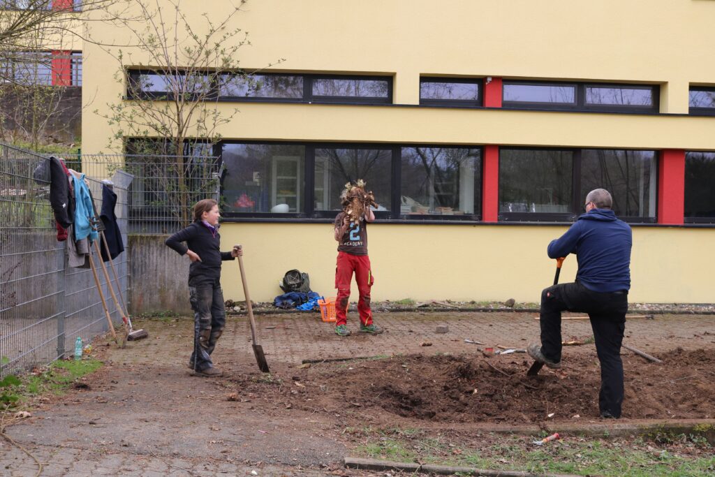 Drei Personen arbeiten im Freien an einem Erdloch neben einem Gebäude.