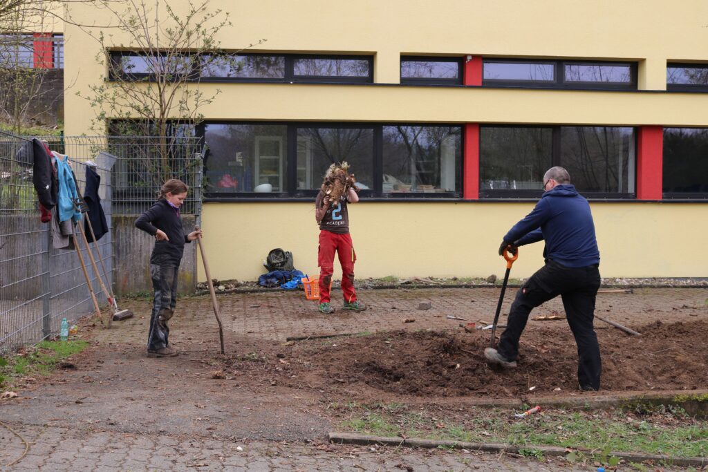 Schüler arbeiten im Schulhof, Stecken im Boden, Gebäude im Hintergrund.