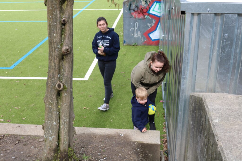 Kinder und Erwachsene auf einem Sportplatz, Baum im Vordergrund.