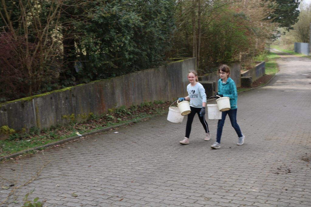 Zwei Mädchen gehen mit Eimern auf einem Weg zwischen Bäumen.