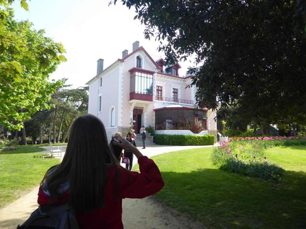 Frau mit Kamera fotografiert eine Villa umgeben von Bäumen und Blumen.
