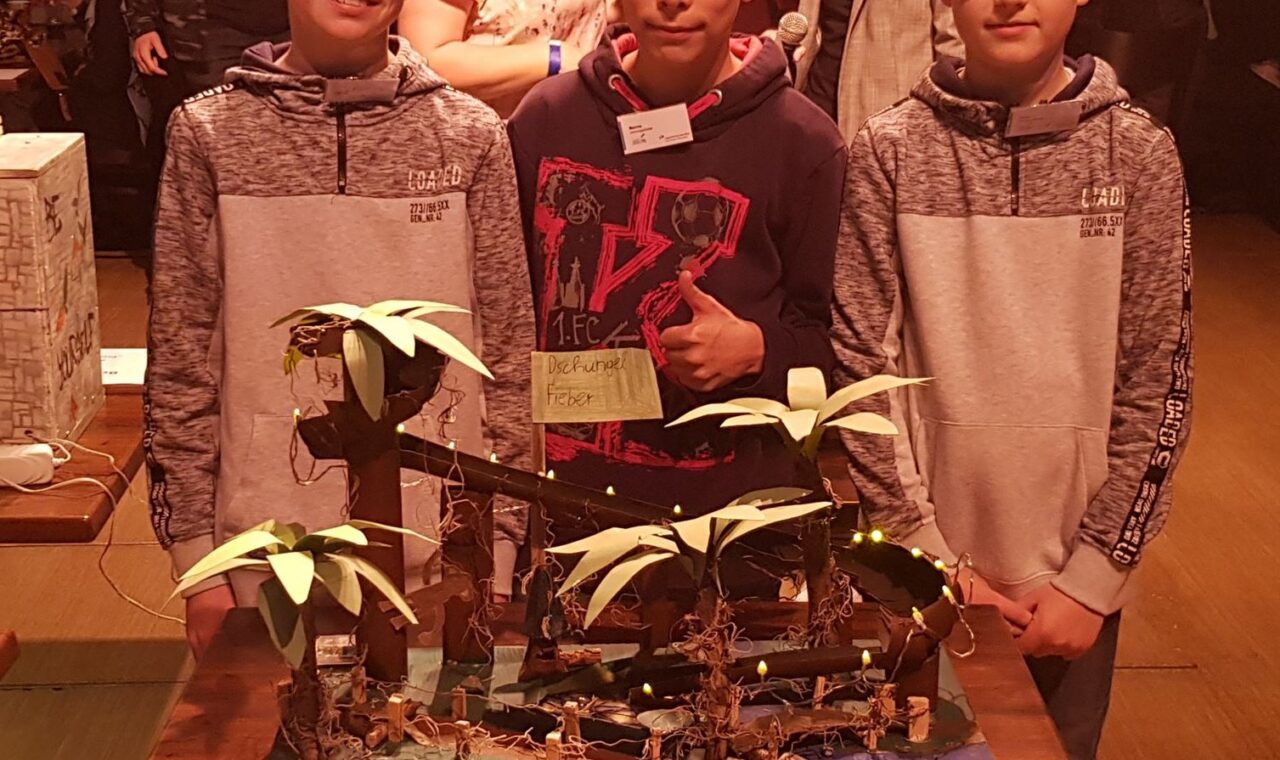 Drei Schüler stehen neben einem Modell mit Palmen und einer Tafel in einem Veranstaltungssaal.