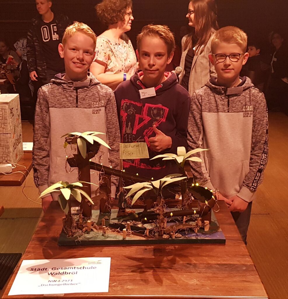 Drei Schüler stehen neben einem Modell mit Palmen und einer Tafel in einem Veranstaltungssaal.