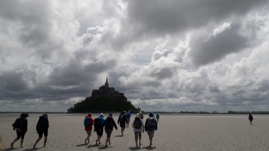 Schüler gehen über einen Sandstrand in Richtung Mont Saint-Michel bei bedecktem Himmel.