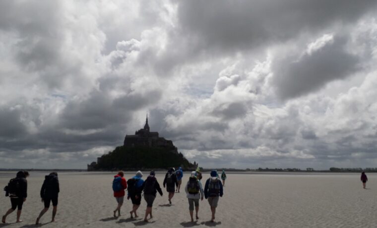 Schüler gehen über einen Sandstrand in Richtung Mont Saint-Michel bei bedecktem Himmel.