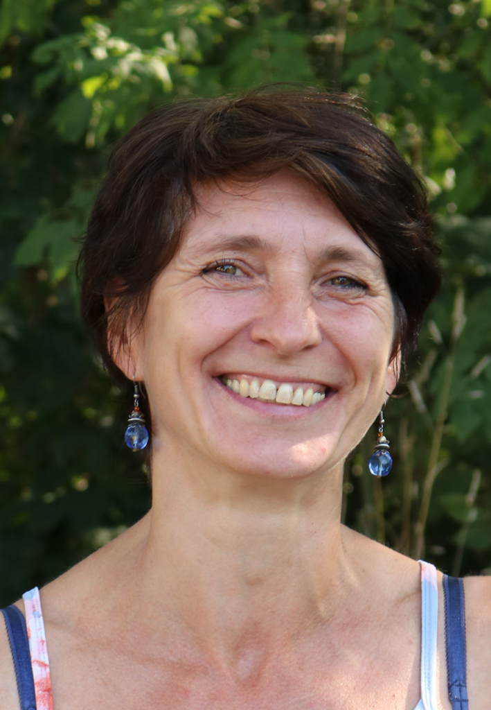Lächelnde Frau mit kurzen Haaren und Ohrringen vor grüner Hintergrund.