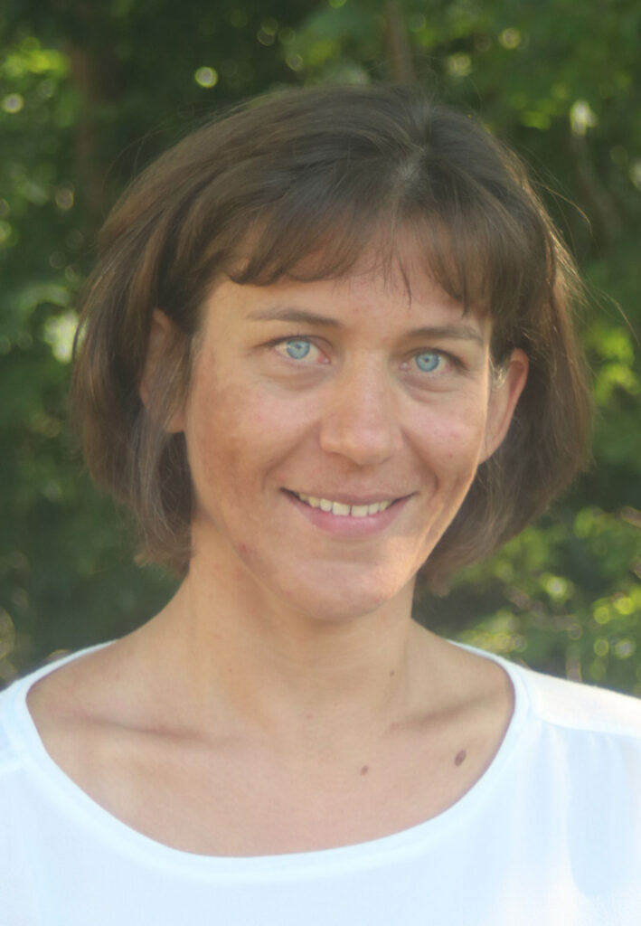 Frau mit kurzen, braunen Haaren und blauen Augen lächelt vor grünem Hintergrund.