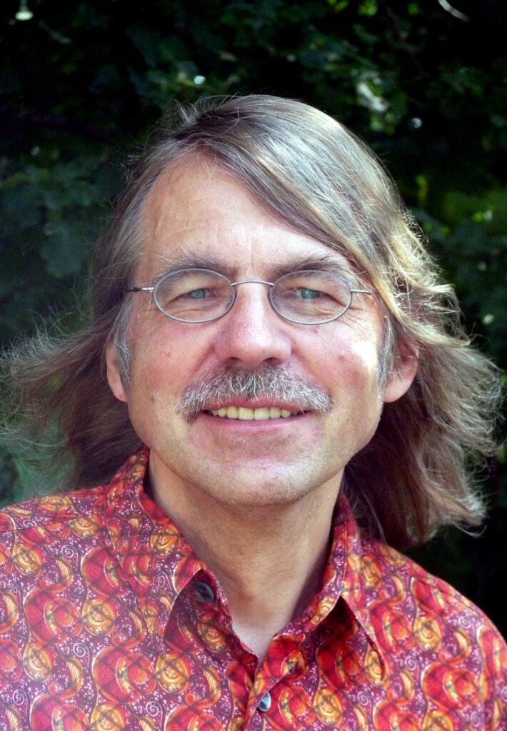 Porträt eines Mannes mit Brille und langem Haar in einem bunten Hemd.