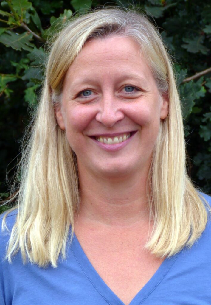 Frau mit blonden Haaren und blauem Shirt lächelt vor grünem Hintergrund.