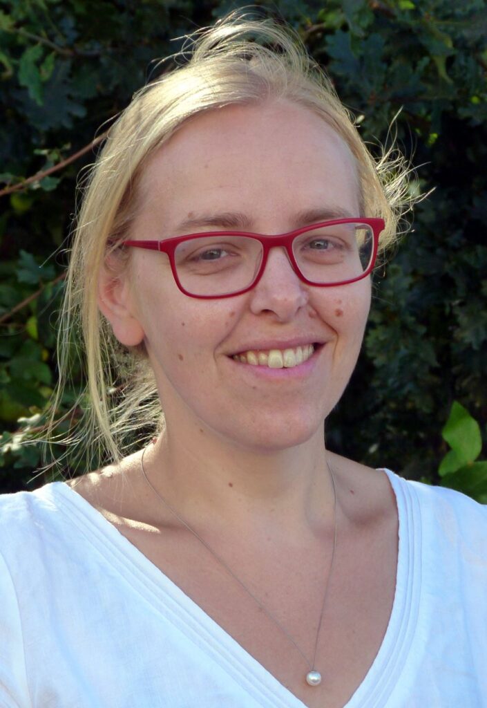 Eine lächelnde Person mit blonden Haaren und roten Brillengestell.