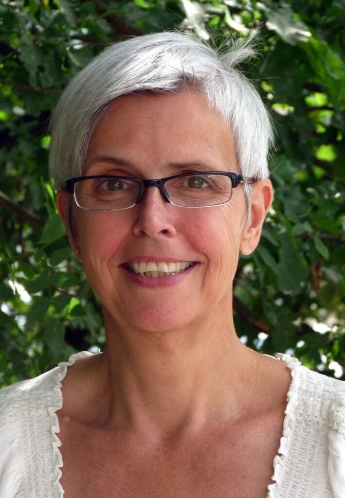 Eine lächelnde Frau mit kurzen grauen Haaren und Brille vor grünem Hintergrund.