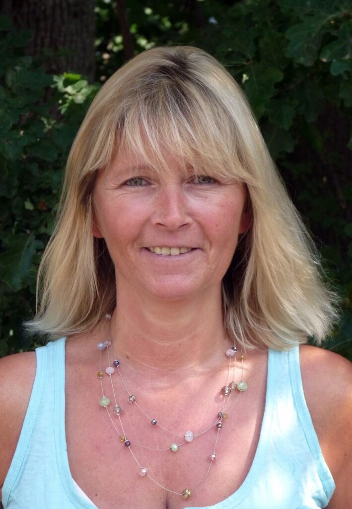 Frau mit blonden, langen Haaren und einem lächelnden Gesicht vor einer grünen Hintergrundkulisse.