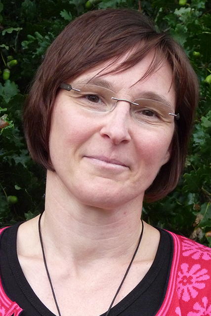 Frau mit kurzem Haar und Brille, lächelt vor grünem Hintergrund.