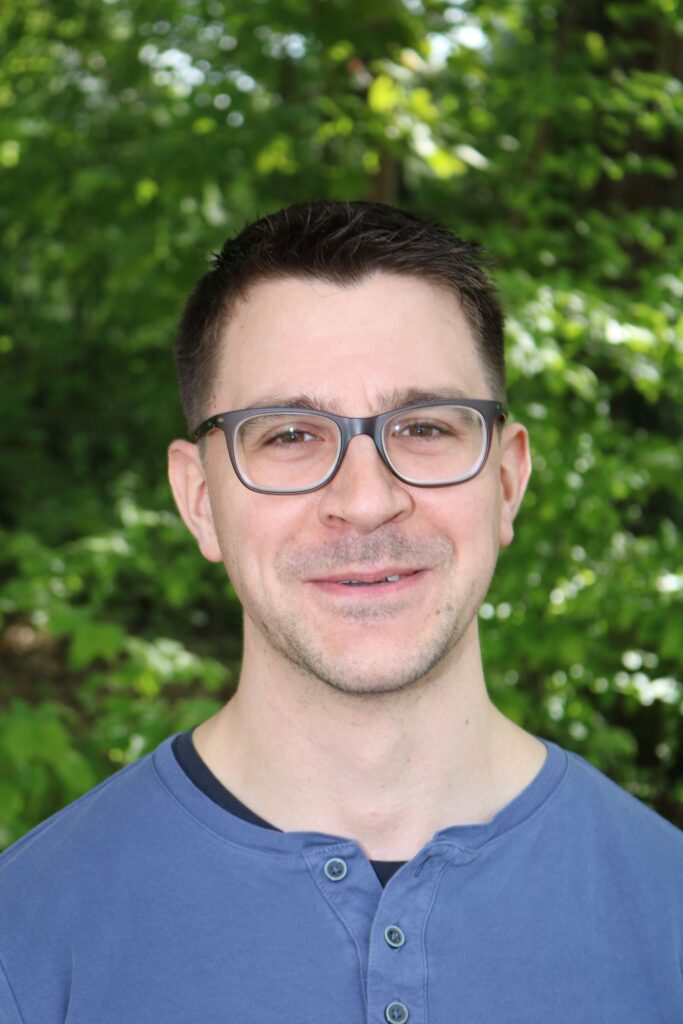 Junger Mann mit Brille vor grünen Pflanzen im Hintergrund.