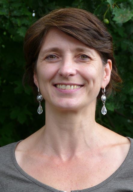 Frau mit kurzen, braunen Haaren und Ohrringen lächelt vor einem grünen Hintergrund.