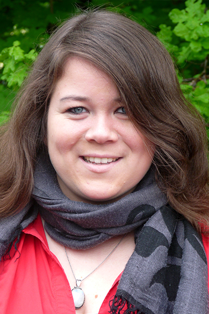 Frau mit langen, dunklen Haaren und Schal vor grüner Pflanzenkulisse.