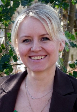 Frau mit blonden Haaren und blauen Augen lächelt vor grüner Hintergrundpflanze.