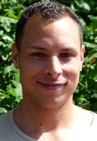 Ein junger Mann lächelt vor einem grünen Pflanzenhintergrund.