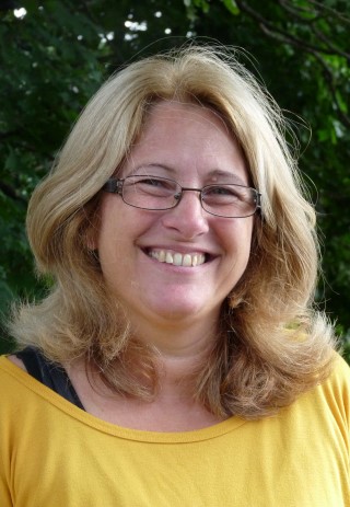 Porträt einer lächelnden Frau mit blonden Haaren und Brille, im Freien.