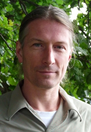 Ein Mann mit langen Haaren steht vor grünen Blättern.