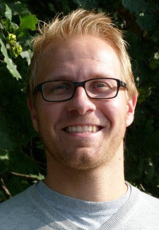 Mann mit Brille und blonden Haaren lächelt vor grünen Pflanzen.