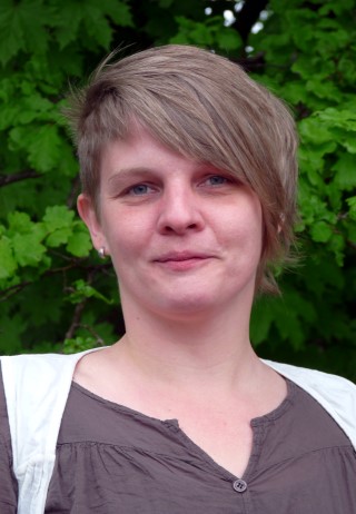 Eine Person mit kurzen Haaren steht vor einem Hintergrund aus grünen Blättern.