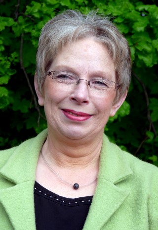 Eine lächelnde Frau mit kurzen, grauen Haaren und Brille vor grüner Vegetation.