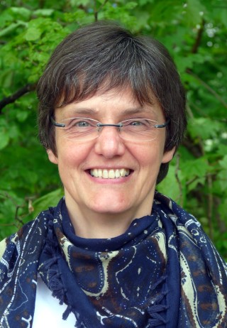 Frau mit kurzer Frisur und Brille, lächelt vor grünem Hintergrund.
