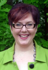Frau mit kürzeren, rötlich-braunen Haaren und Brille vor grüner Pflanzenkulisse.