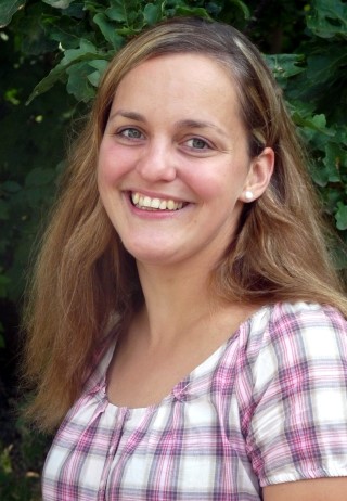 Frau mit langen Haaren in kariertem Oberteil lächelt vor grüner Kulisse.