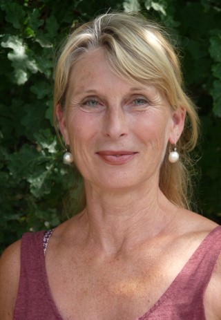 Frau mit blonden Haaren und Perlenohrringen vor grüner Hintergrundpflanze.