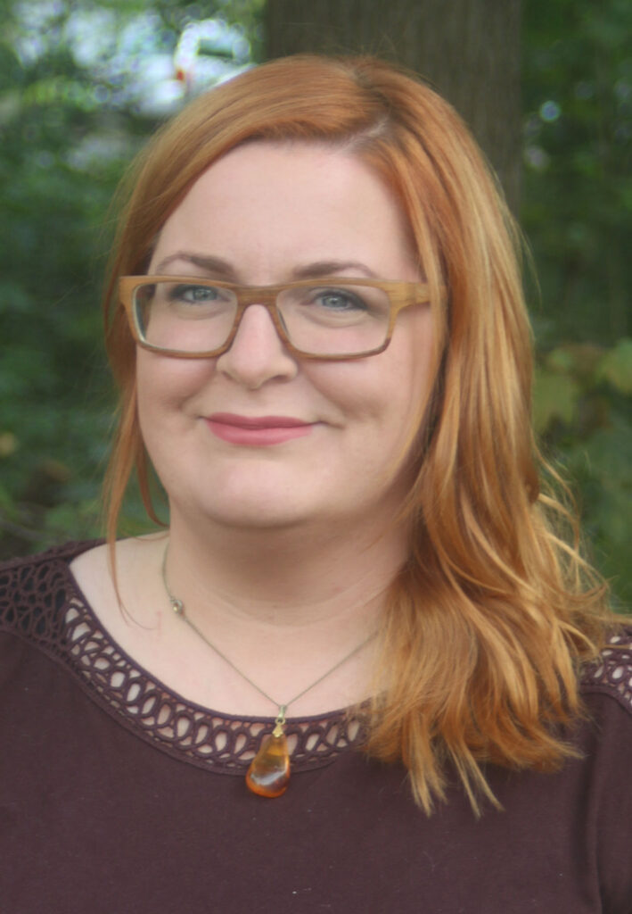 Frau mit roten Haaren, Brille und einer Kette steht im Freien vor unscharfem Hintergrund.
