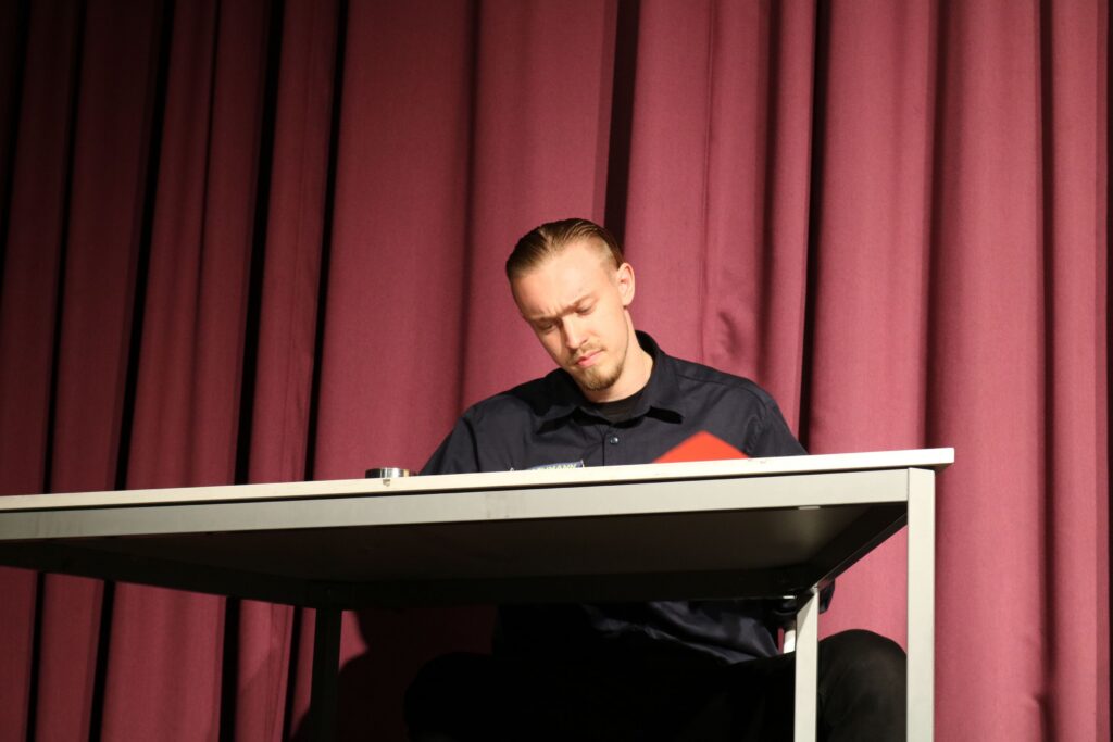 Ein Mann sitzt an einem Tisch und schreibt, hinter ihm ist ein roter Vorhang.
