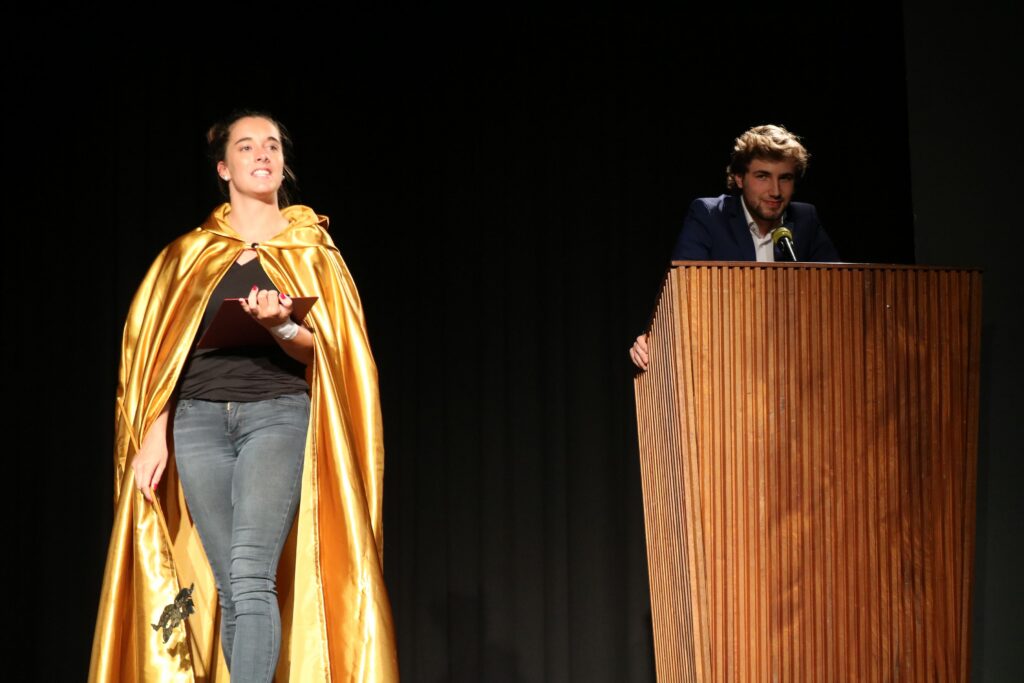 Eine Person mit goldener Umhüllung steht neben einem Rednerpult.