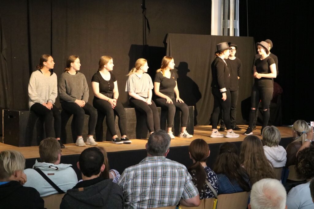 Theateraufführung mit Schauspielern auf der Bühne und Publikum im Vordergrund.