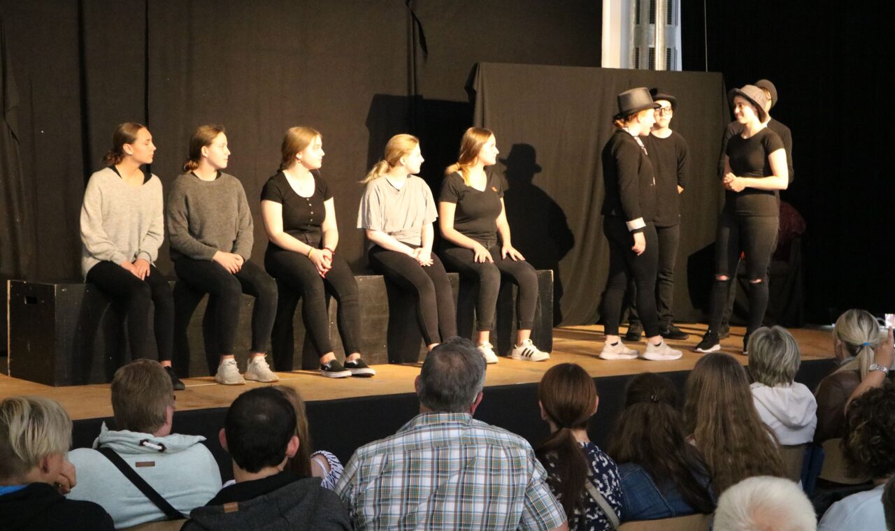 Theateraufführung mit Schauspielern auf der Bühne und Publikum im Vordergrund.