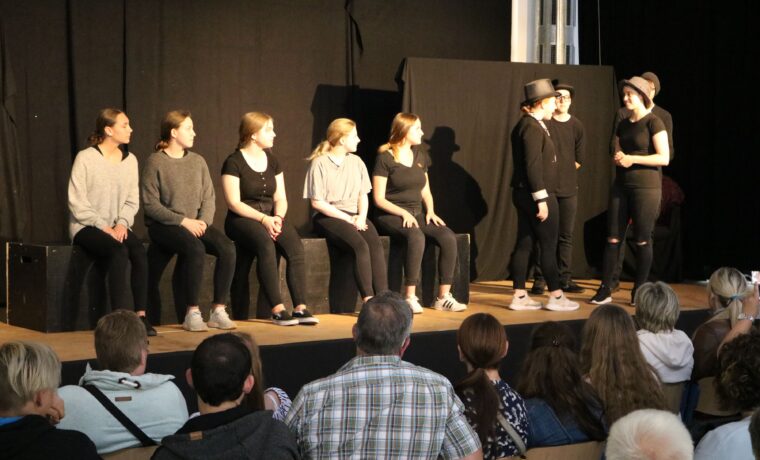 Theateraufführung mit Schauspielern auf der Bühne und Publikum im Vordergrund.