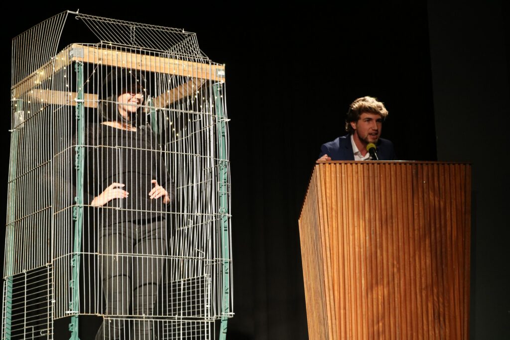 Ein Mann spricht am Podium, während eine Person in einem Käfig steht.
