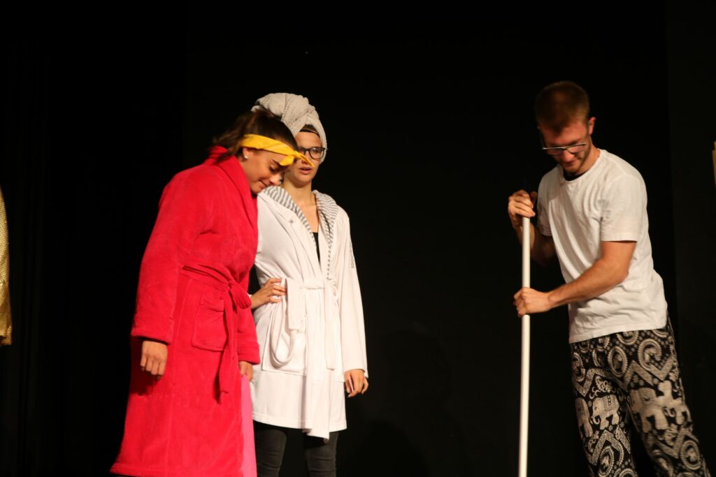 Drei Schauspieler in Kostümen auf einer Bühne, zwei schauen interessiert zu.
